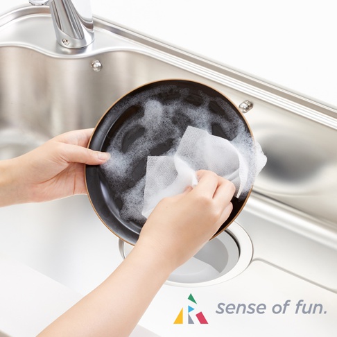 雙手拿著一個黑色帶有棕色邊緣的盤子，盤子裡有泡沫。一隻手用一張白色、網格狀的布擦拭盤子，另一隻手扶著盤子。背景是閃亮的銀色水槽和水龍頭。