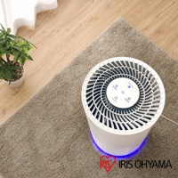 白色圓柱形空氣清淨機，頂部有放射狀的白色扇葉和藍色指示燈，底部散發著藍光，置於米色絨毛地毯上。