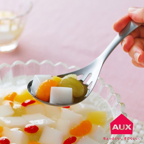 一只手拿著一支銀色金屬濾水菜匙，匙中盛有塊狀白色果凍、橘色柑橘片、綠色葡萄及其他水果丁，正從一碗裝滿果凍和水果的透明玻璃碗中舀取。