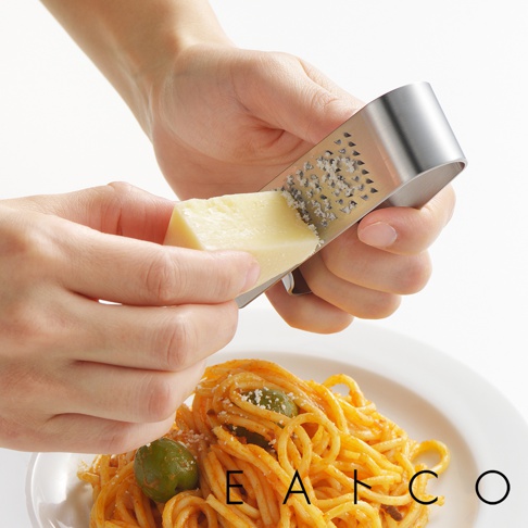 一隻手持銀色不鏽鋼立式磨泥器，另一隻手將一塊淡黃色起司在磨泥器上摩擦，磨出細屑。下方是盤中擺放的義大利麵。