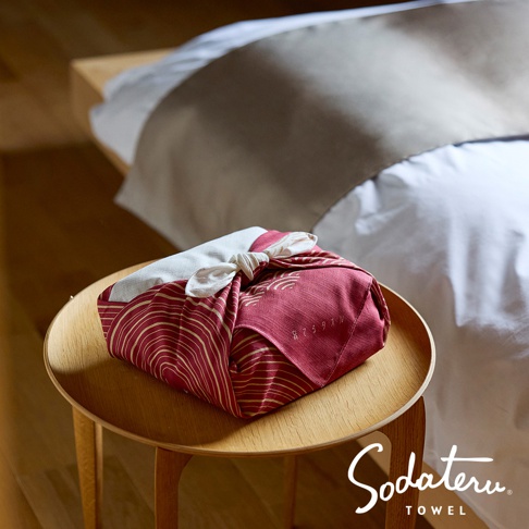 由「Sodateru TOWEL」品牌標誌標註的浴巾，以日式風呂敷（furoshiki）包裝包裹著，呈現在木製小圓桌上，背景是整潔的床鋪。