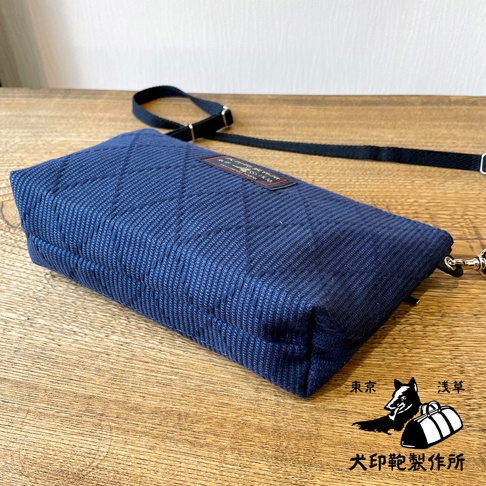 深藍色刺子織隨身斜背包，配有黑色可調式斜背帶和銀色金屬扣環。包包表面有菱格紋縫線，正面中央有一個紅色標籤。