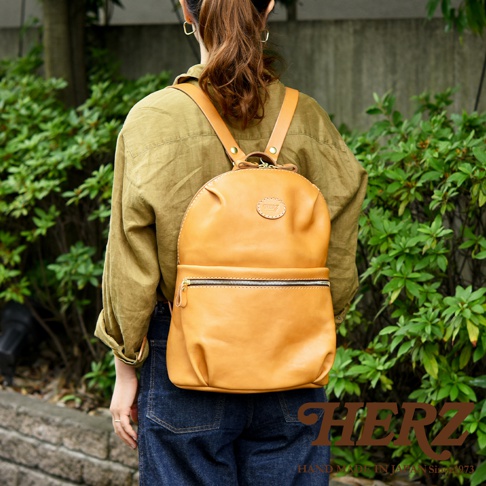 駝色皮革大開口圓角後背包，配有拉鍊前袋和圓形品牌標籤，由品牌 HERZ 製造。