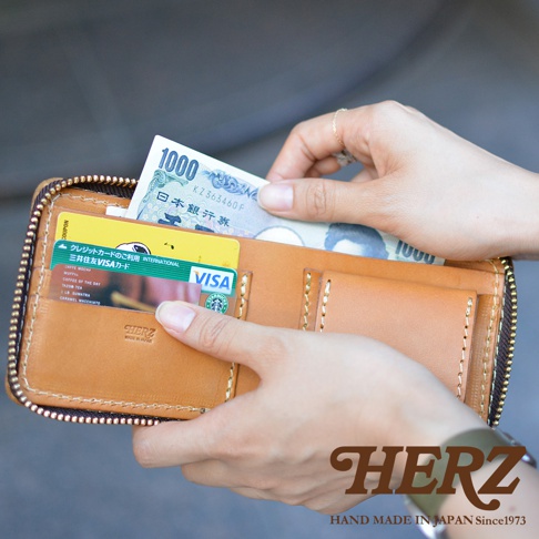 一位女性手持日本HERZ品牌的棕色手工皮革拉鍊短夾，皮夾內插有一張星巴克隨行卡及三井住友VISA信用卡，同時正在放入一張一千元日幣紙鈔。皮夾呈現經典的皮革縫線細節與簡約職人質感，右下角印有HERZ品牌名稱與相關標語。