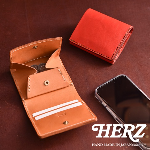 一張深棕色桌面上展示著 HERZ 品牌的手工皮革短夾。畫面中一個亮橘紅色的迷你短夾呈摺疊狀態，旁邊則有一個展開的淺棕色短夾，內部設計有硬幣袋與兩個卡片插槽，並露出少量硬幣。右下角放置著一支裝在透明手機殼中的智慧型手機，整體呈現職人手作的質感與簡約風格。