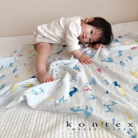 一嬰兒趴蓋在印有可愛動物塗鴉圖案的白色二層紗嬰兒毯上。毯子上有藍色、粉色和黃色的恐龍、鳥類和長頸鹿等圖案。