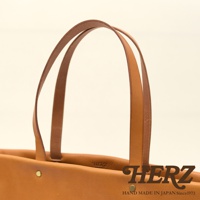 職人手作皮革極簡托特包，採用天然皮革材質，呈現溫潤的焦糖棕色。包身結構簡潔，配有兩個皮質提把，提把與包身以金屬鉚釘固定，質感樸實。