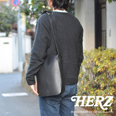 日本人身揹黑色皮革直式斜背包，搭配深灰色毛衣與藍色牛仔褲，包包上有HERZ品牌標誌，圖片底部標示「HERZ HAND MADE IN JAPAN Since1973」。