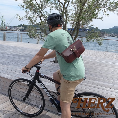 斜挎著一個棕色皮革單扣斜背包的男子正在騎自行車，背包上有一個金屬扣環。