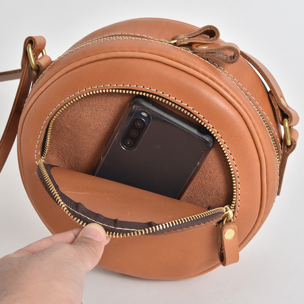 棕色皮革製成的手作小圓包，包身圓潤，拉鍊開口處露出手機，展現其收納空間。