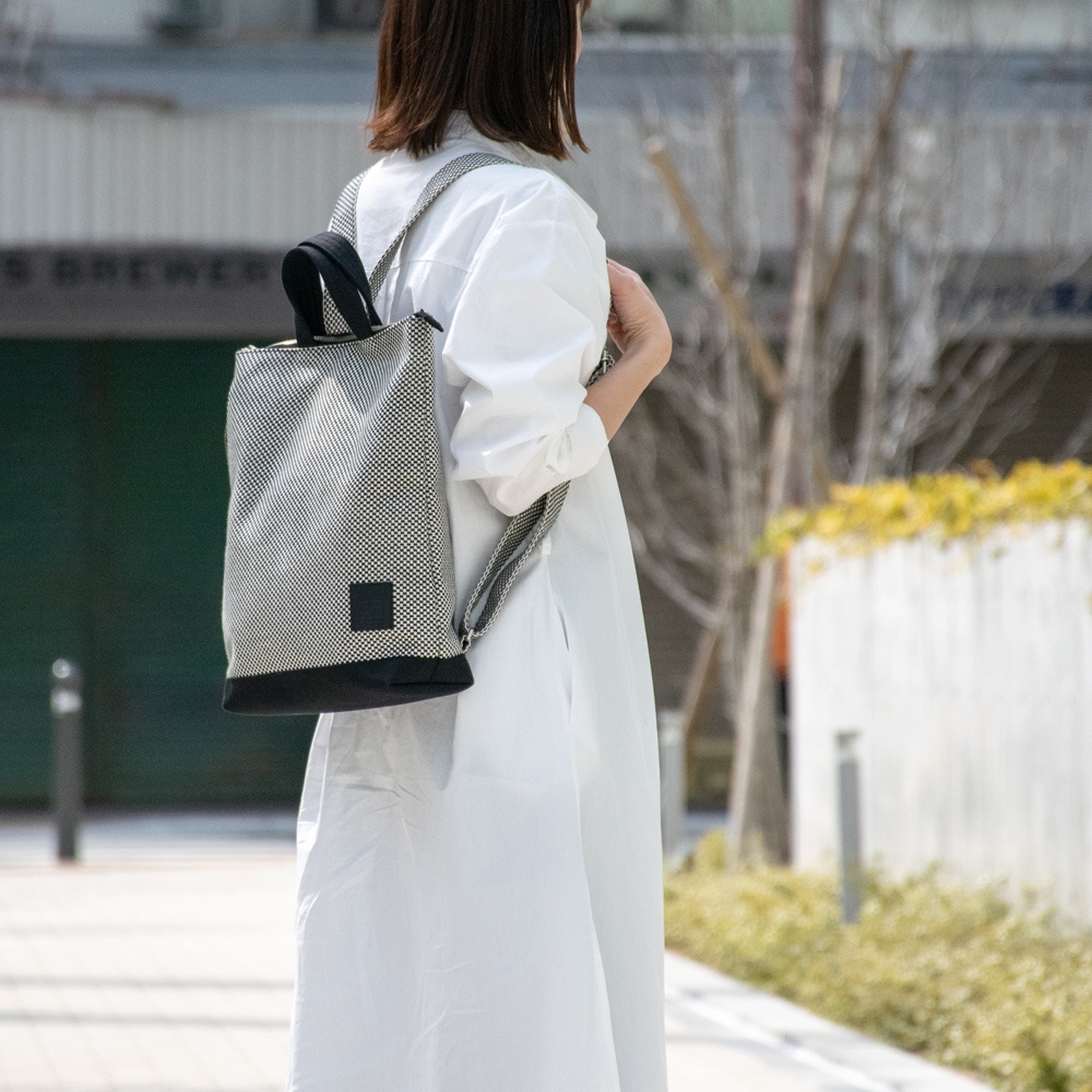 女性背著一個以三河木綿刺子織製成的手提後背包，背包主體為灰白色網格圖案，底部為黑色，側面有一個黑色方形標籤。