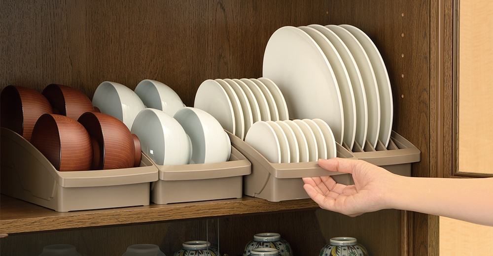 櫥櫃內擺放著兩個淺褐色餐盤收納架，左側架上收納著木質碗，右側架上則收納著白色盤子，一只手正在將右側的收納架往外拉。