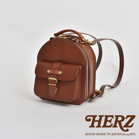 職人手作咖啡色皮革迷你後背包，圓潤造型，正面有翻蓋口袋與金屬扣環，側邊有拉鍊，附有皮革揹帶。