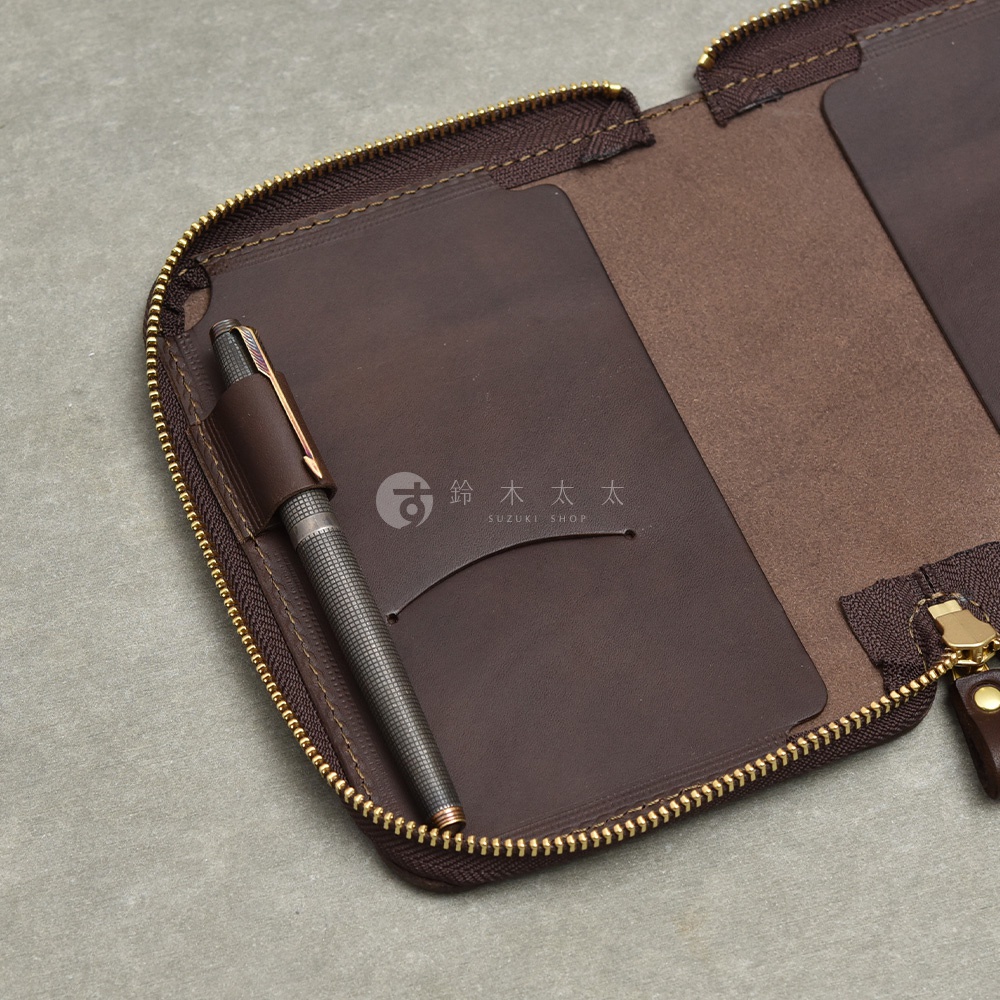 職人手作皮革Organ記事本收納包，深棕色皮革內側有一個筆套，筆套內插著一支銀色格紋金屬筆，收納包採用金色拉鍊設計，內部有卡片插槽。