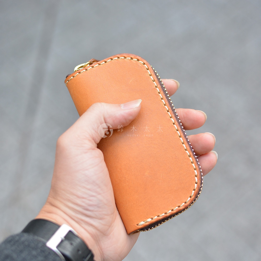 一只手正拿著一個棕褐色皮革製的橢圓形拉鍊鑰匙包，包包有著縫線細節和金屬拉鍊。