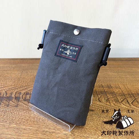 犬印鞄製作所純綿10號帆布手機包，灰黑色，附有品牌標籤與掛繩細節，置於木質桌面上的透明支架上。