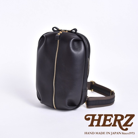 HERZ 品牌立體黑色皮革斜背包，配有金色拉鍊和品牌標誌。