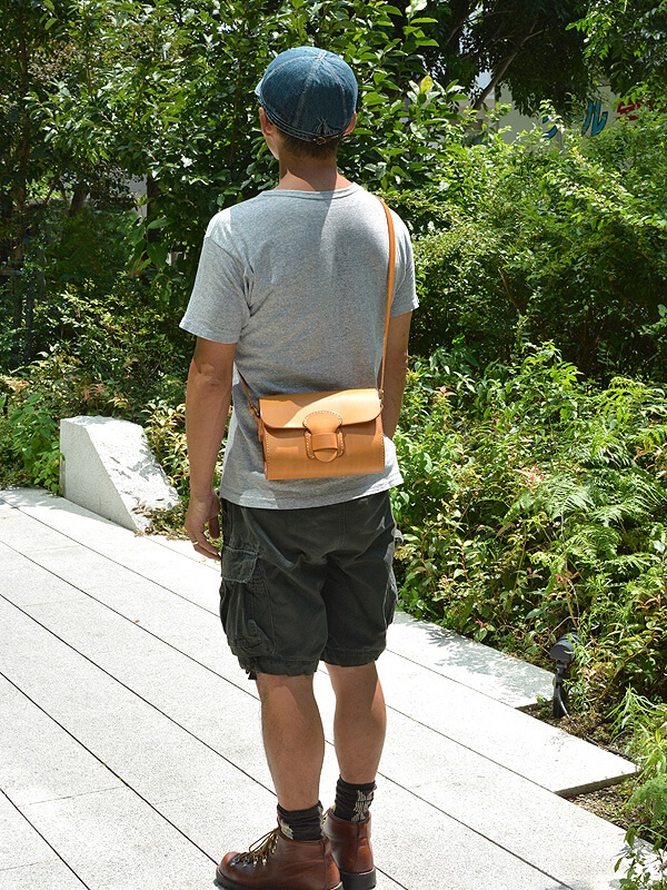 一位男性身穿灰色T恤、卡其色短褲，斜挎著一個淺棕色皮革馬鞍包，包上有寬版皮革扣帶。