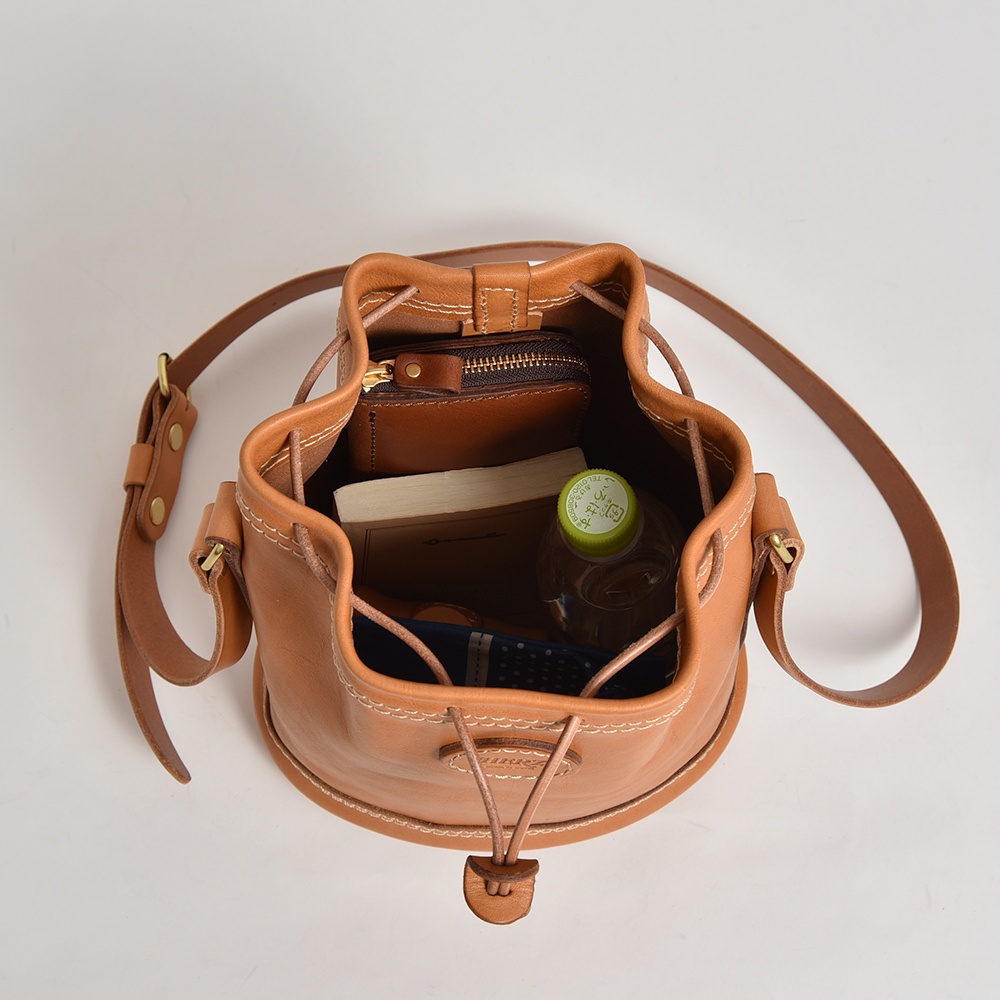 職人手作皮革束口單肩包，原色皮革材質，包身束口設計，內部可見錢包、書本、水瓶及印花布飾。