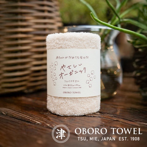 一條淺米色、織紋精緻的有機棉毛巾，標籤上印有日文文字與可愛的棉花圖案，標示「OBORO TOWEL」。