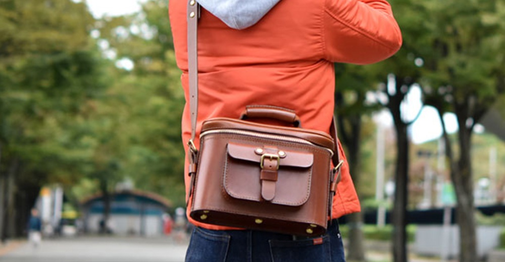 一個身穿橘色外套的人斜揹著一個棕色皮革相機包，包上有一個掀蓋式口袋和金屬扣。
