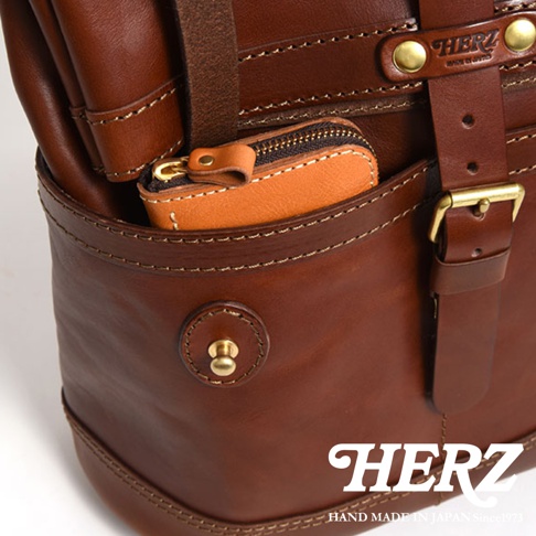 職人手作皮革雙口袋折蓋後背包 R-301，深棕色皮革，設有雙前口袋，其中一個口袋內放有一個小巧的橘棕色皮革零錢包。包款採用縫線裝飾，搭配金屬釦環和鉚釘，展現質感。