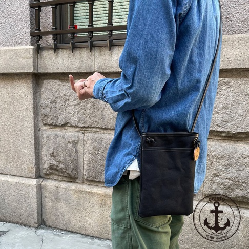 黑色帆布皮革散步包，配有可調節肩帶和外部口袋。包包上有一個圓形吊牌，上面印有船錨圖案和「clueto KOBE since 2001」字樣。