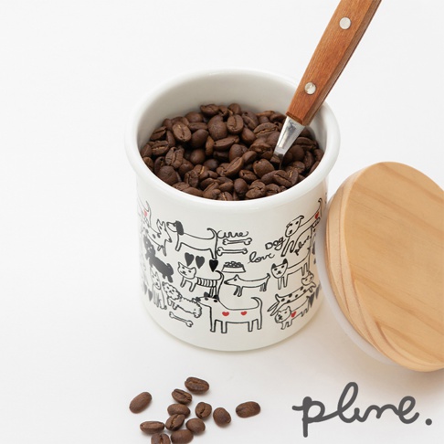 白色琺瑯儲物罐，外觀印有黑色線條描繪的各式狗狗圖案，罐內裝有咖啡豆，一把木柄金屬勺插入罐中。旁邊擺放著一個木質圓形蓋子，以及幾顆散落的咖啡豆。