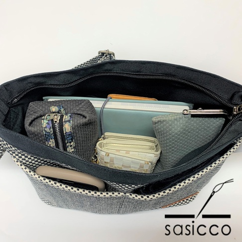 三河木綿刺子織肩背包，外觀為黑白格紋，內部分層收納，可見灰色筆袋、淺色編織錢包、藍色筆記本及手機。