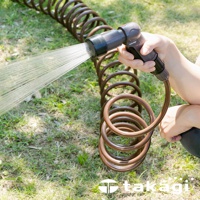 一隻手正握著一個灰色和黑色的灑水噴頭，噴出細長的水柱。噴頭連接到一條棕色的螺旋狀水管，水管鋪設在綠色的草地上。