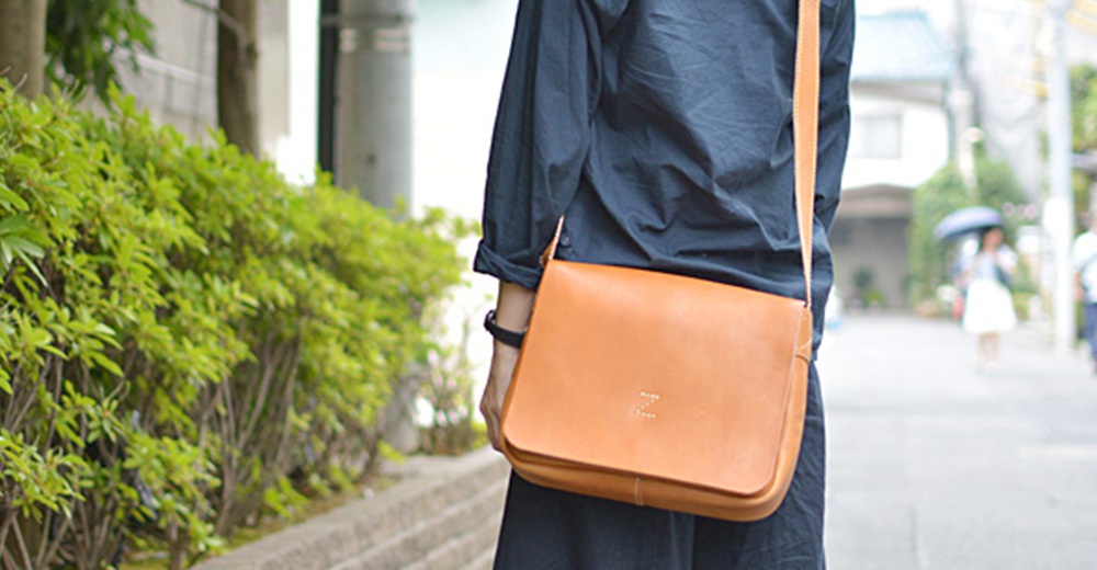 一位身穿深藍色襯衫的人背著一個皮革製的駝色側背包，走在綠色植物和街道旁