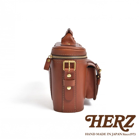 HERZ 品牌標誌，下方有 HAND MADE IN JAPAN Since1973 字樣，圖片主體為一個棕色皮革製成的圓筒狀相機包，採用側邊視角拍攝，包包帶有拉鍊、金屬釦環及側邊小口袋等細節。