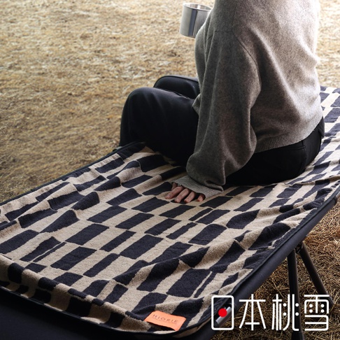一位身穿灰色毛衣和黑色褲子的人坐在一張露營椅上，腿上鋪著一條帶有深藍色和米色格子圖案的毛巾，毛巾角落處有「HIORIE」標籤，右下角有「日本桃雪」的文字標記。