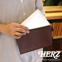職人手作皮革三角底拉鍊手拿包，深棕色皮革，金色拉鍊，包面上壓印有「HERZ」品牌標誌。