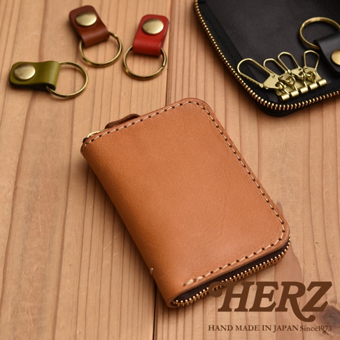 職人手作皮革多用途收納鑰匙包，主體為駝色皮革，配有金色拉鍊和四個金屬鑰匙扣。旁邊展示有綠色、紅色和棕色的皮革鑰匙圈。