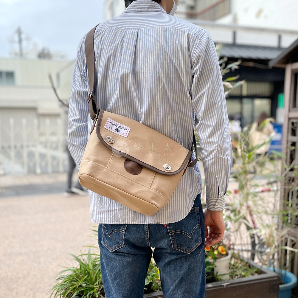 淺褐色純綿帆布製成的雙扣郵差包，帶有咖啡色皮革飾邊和標籤，斜背於身穿藍白條紋襯衫的男性身上。