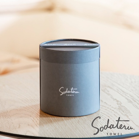 灰色圓筒形包裝盒，上面印有白色「Sodateru TOWEL」字樣，盒蓋上有一條裝飾性銀色緞帶，緞帶上重複印有「Good」字樣。