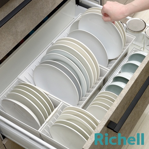 抽屜式餐盤收納架整齊擺放著白色及米白色系的各式盤子，一隻手正在從收納架中拿取盤子，收納架旁還有擺放碗的空間。