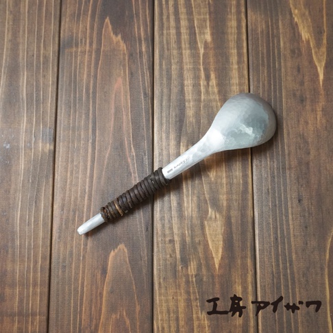 銀色鋁製湯勺，勺頭呈圓弧狀，表面有錘目紋理。勺柄部分以棕色皮革纏繞，末端為銀色尖頭。