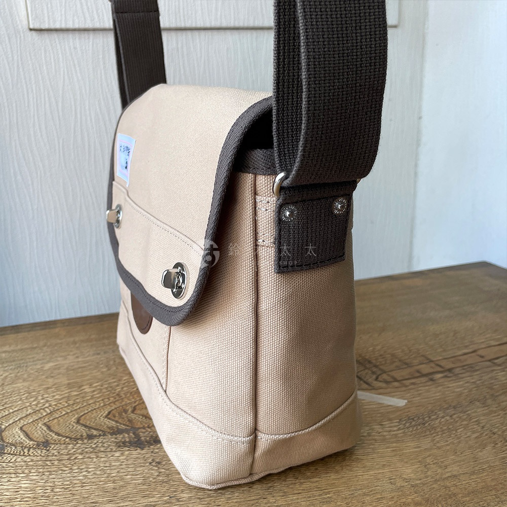 犬印純綿帆布製的米色雙扣郵差包，配有深棕色帆布肩帶和邊緣，包蓋上有銀色金屬旋扣和一個小方形貼布。