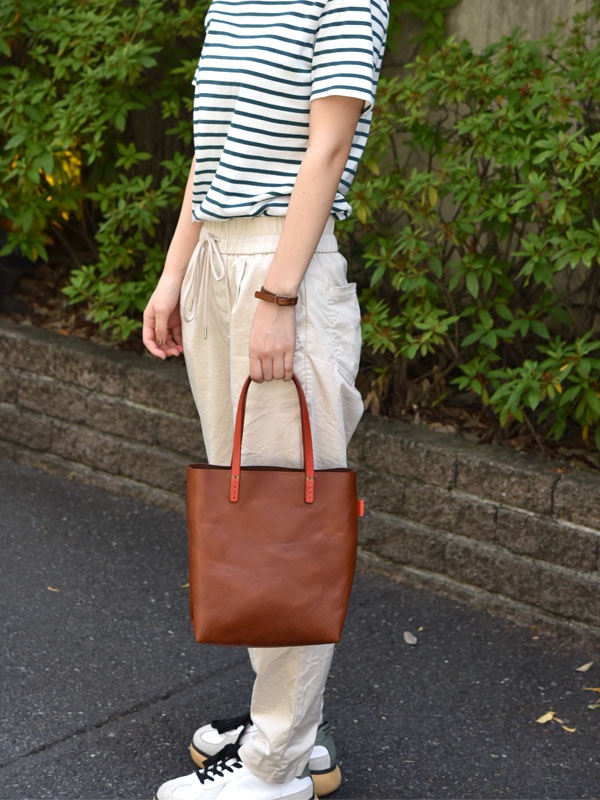 棕色皮革直式軟皮托特包，配有橘色提把，包側有橘色品牌標籤，由一位身穿條紋上衣、米色長褲的女性持握。