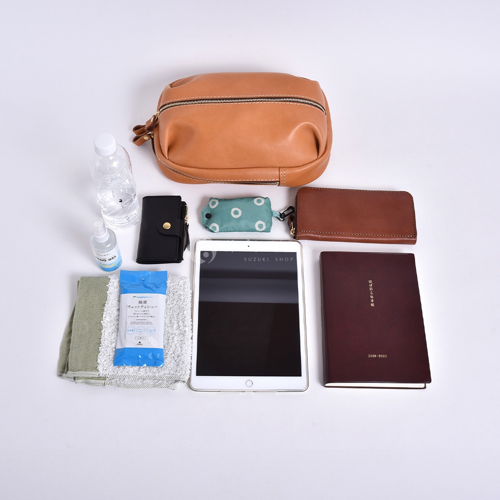 駝色皮革運動斜背包，內含一瓶水、一個黑色皮夾、一個綠色圓點圖案的收納包、一個平板電腦、一本深紅色筆記本、一塊毛巾和一包濕紙巾。