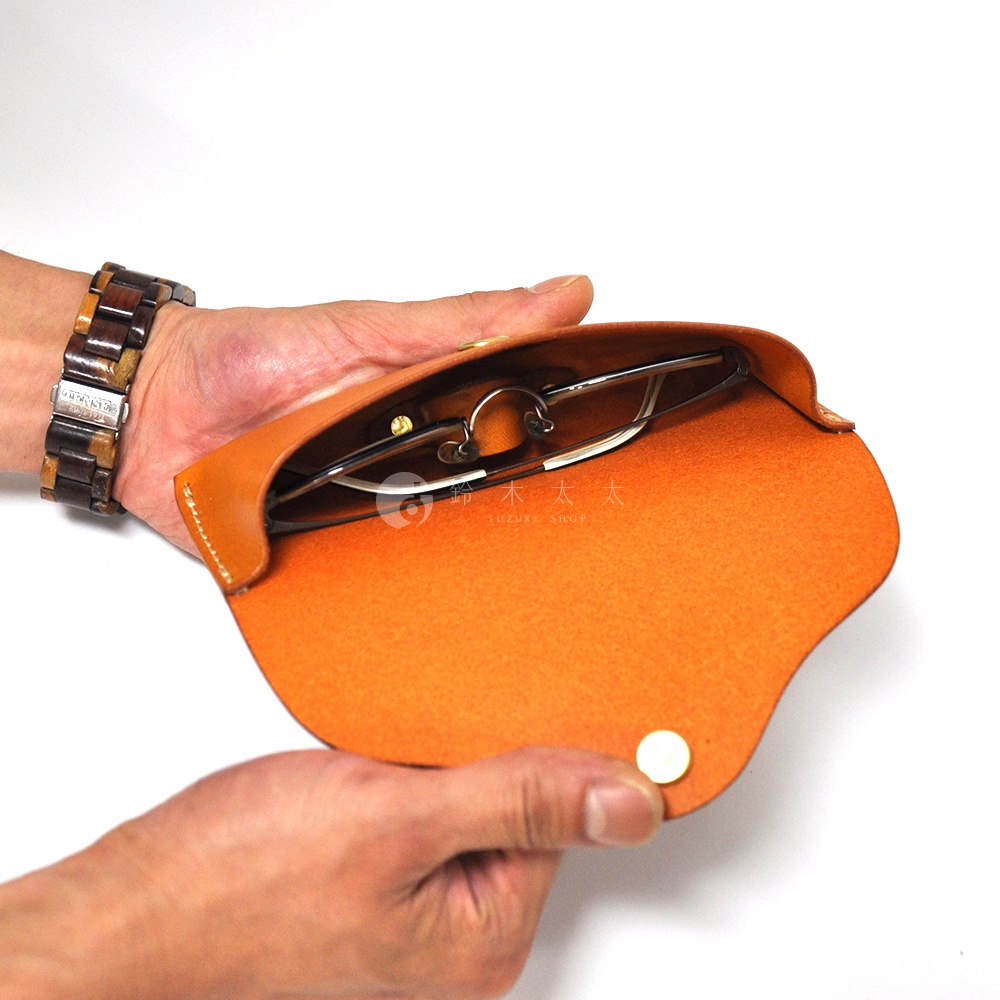 一雙手拿著橘色皮革眼鏡套，裡面放著一副金屬細框眼鏡，手腕上戴著木質手鍊。