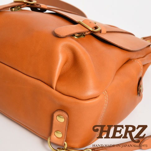 這是一款由日本職人手工製作的HERZ品牌經典皮革後背包，採用棕色牛皮打造，展示了包身側邊與頂部金屬扣環的特寫細節。包體線條圓潤且皮革質感厚實，並配有堅固的黃銅鉚釘與精緻的車縫線，底部印有HERZ品牌商標與手作標誌。