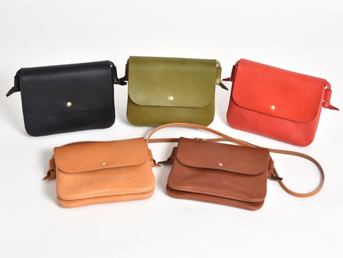 五款職人手作皮革橫式隨行袋，顏色分別為黑色、橄欖綠、紅色、淺棕色和深棕色，皆為方形翻蓋設計，附有金色金屬釦和皮革背帶。