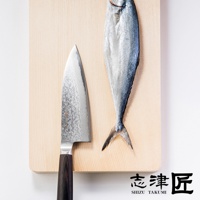 一把銀色金屬材質、帶有紋理的 Deba 魚肉料理刀，刀柄為深棕色，與一條銀灰色處理過的魚並列於木質砧板上。