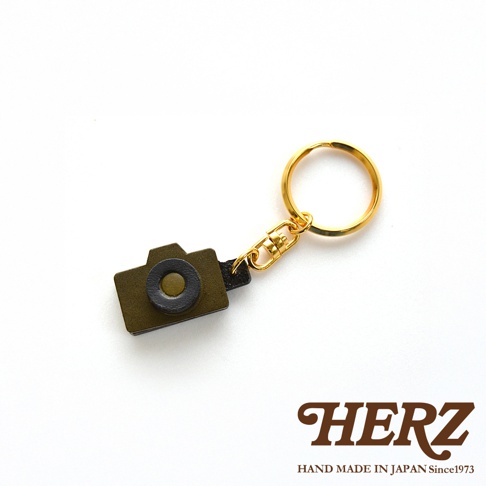 職人手作皮革相機造型鑰匙圈，採用橄欖綠皮革製成，搭配黑色鏡頭裝飾，配有金色鑰匙圈與連接環。