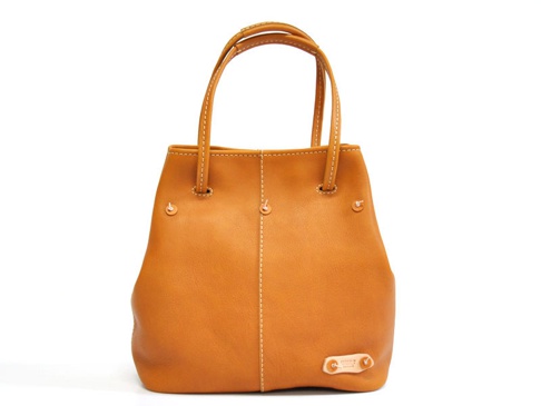 焦糖色皮革托特包，有雙提把、縫線裝飾、側邊鉚釘及底部皮革標籤。