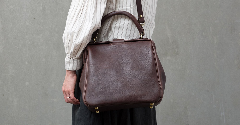 棕色皮革製的方形口金托特包，配有圓形提把和可拆卸肩帶，包包底部有四個金屬腳釘。