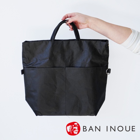 BAN INOUE 品牌標誌旁是一個黑色的亞麻布托特包，由一只手拿著，包包上有兩個前袋和一個可拆卸的肩帶。